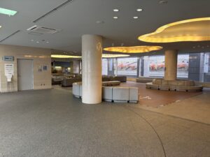 2階共同待合ロビー | 遺産相続手続まごころ代行センター Spacious lobby with circular beige couches arranged around a marble column and warm yellow curved ceiling lights along the ceiling edge.