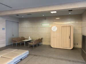 2階共同待合ロビー2 | 遺産相続手続まごころ代行センター Waiting area with beige marble walls, row of brown chairs, and a wooden reception panel with a circular logo reading'mamaro' on the right side.