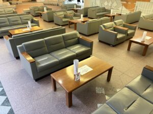 2階共同待合ロビー3 | 遺産相続手続まごころ代行センター Spacious lounge area with many olive-green leather armchairs and wooden tables, menus on the tables, in a public waiting area like an airport or mall.