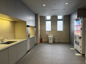 2階共同待合ロビー4 | 遺産相続手続まごころ代行センター Office kitchenette/break room with gray cabinets along left wall, yellow accent wall, and a vending machine on the right; tiled gray floor and overhead lighting.