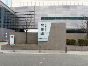 北斎場外観 | 遺産相続手続まごころ代行センター Exterior of a modern grey building with a large pale sign displaying Japanese characters, indicating Osaka city facility and parking entrance nearby.