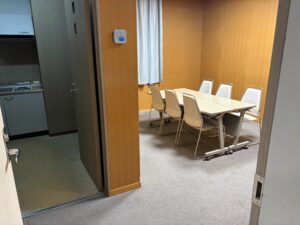 小式場導師控室 | 遺産相続手続まごころ代行センター Conference room with a long table and beige chairs against warm wooden walls; left side shows a kitchen area with appliances partially visible.
